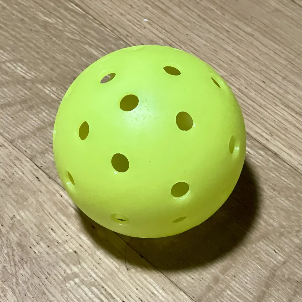 示意照片：一顆螢光黃綠色洞洞球放在淺色木紋地板上，球體右側落下一道柔和陰影